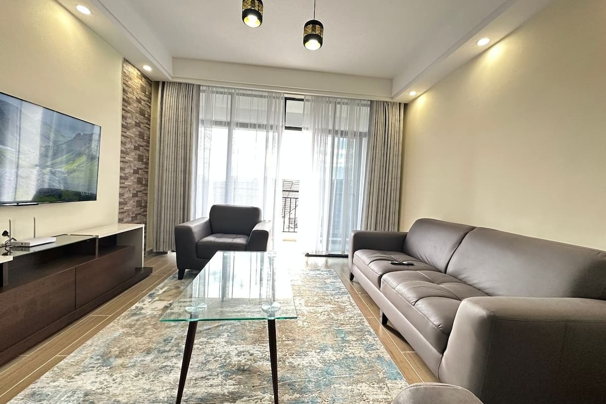 A modern living room with a glass coffee table, leather sofa, and a flat-screen TV mounted on the wall.