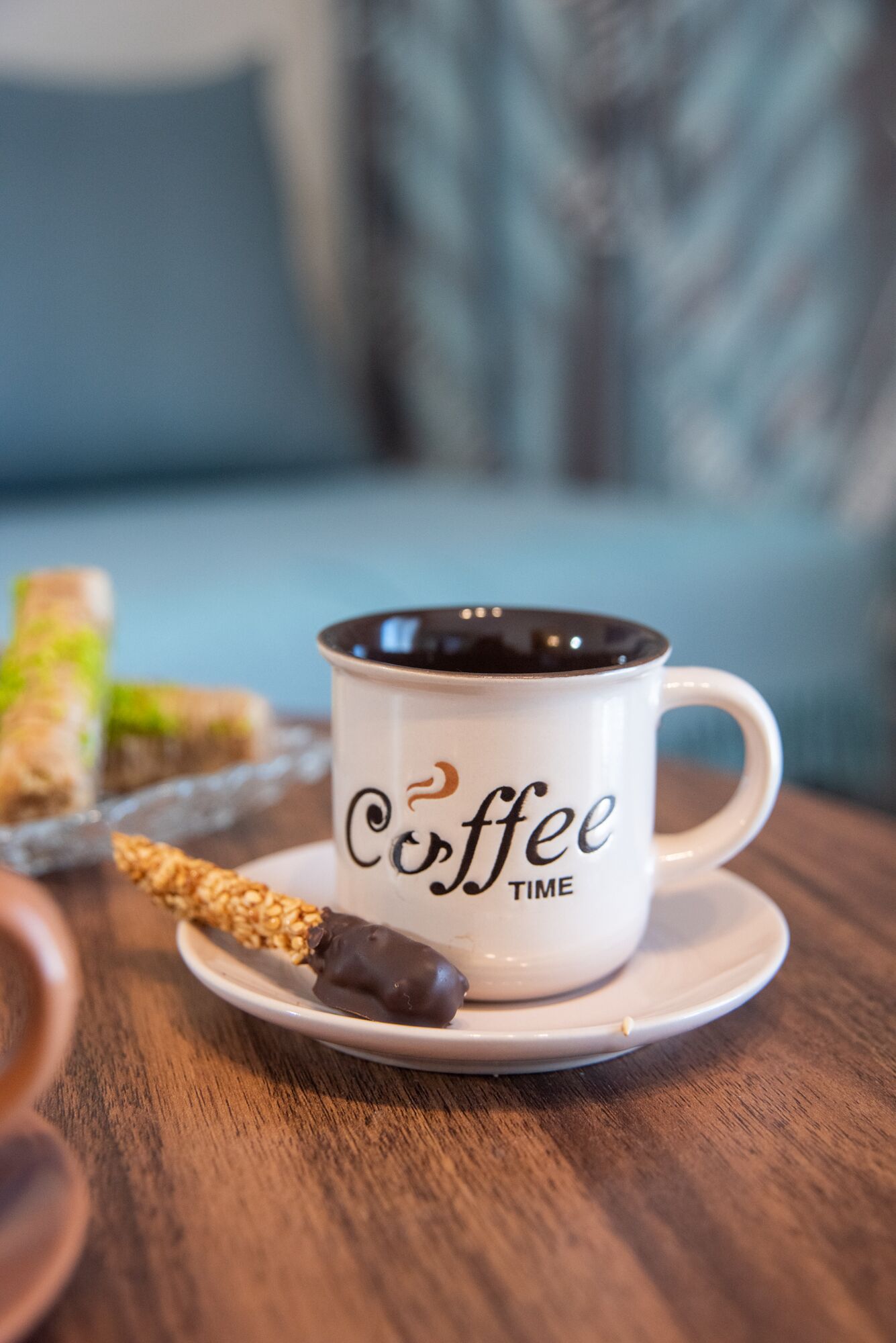 A white coffee mug with "Coffee Time" text on a saucer, a chocolate-covered treat, and a spoon on a wooden table.