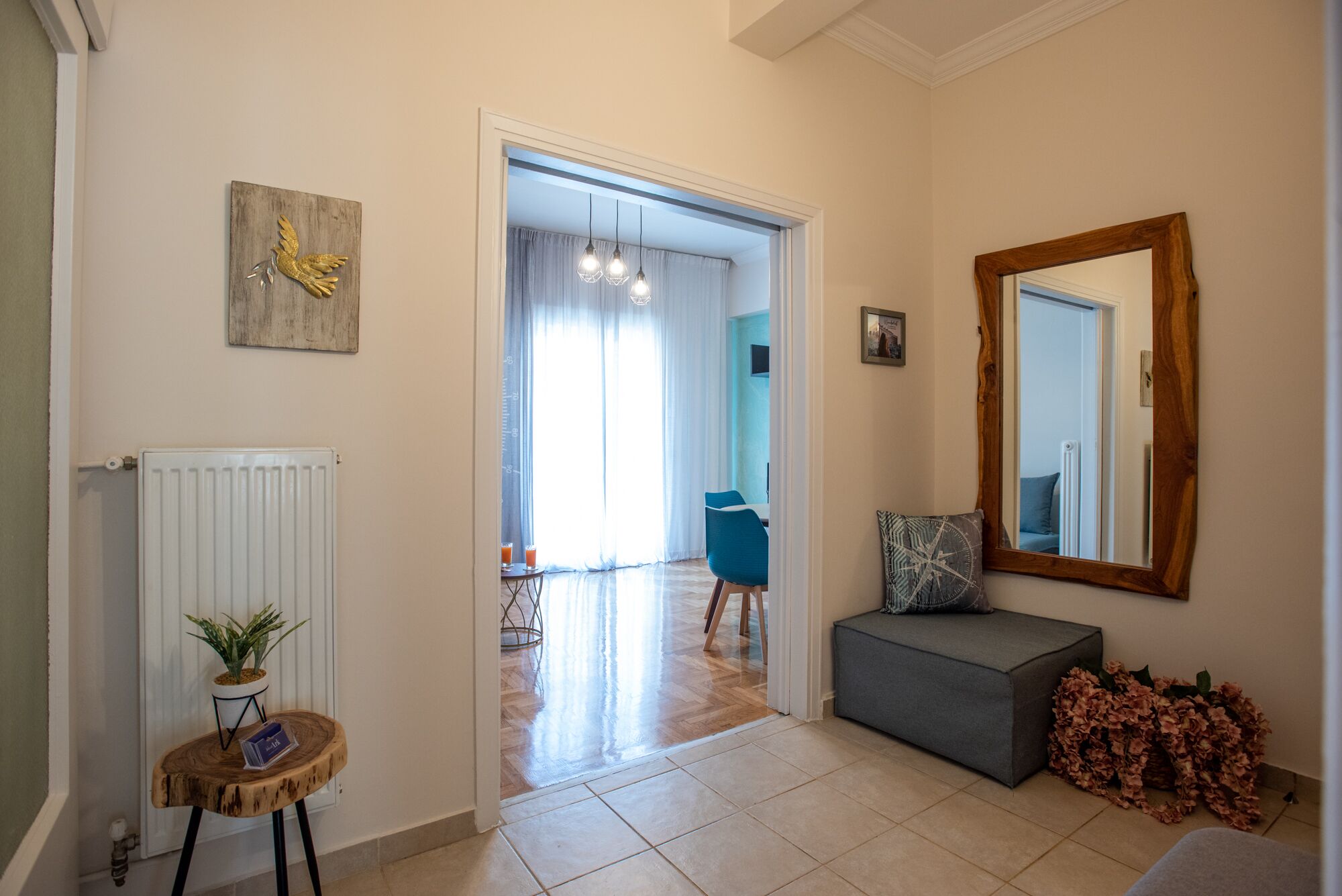 A hallway with a radiator, a wooden stool with a plant, a blue chair, a gray bench, and a mirror on the wall.