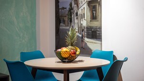 A table with a fruit bowl and two blue chairs in a room with a view of a street through a window.