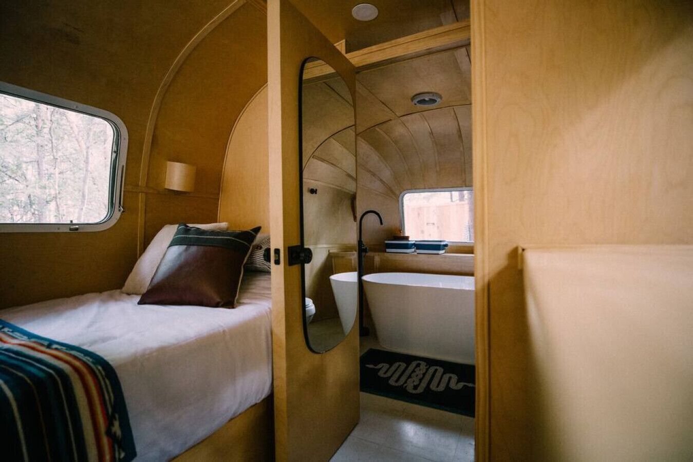 A compact RV interior with a bed, a small bathroom with a bathtub, and a window providing natural light.