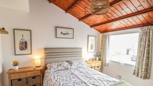 A bedroom with a wooden ceiling, a bed with floral bedding, a bedside table with a lamp, and a window with curtains. - Branwen (Porthmadog)