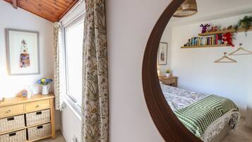 Un dormitorio con techo de madera, una cama con colcha de cuadros verdes y blancos, una mesita de noche con lámpara, un aparador con cestas y una ventana con cortinas florales.