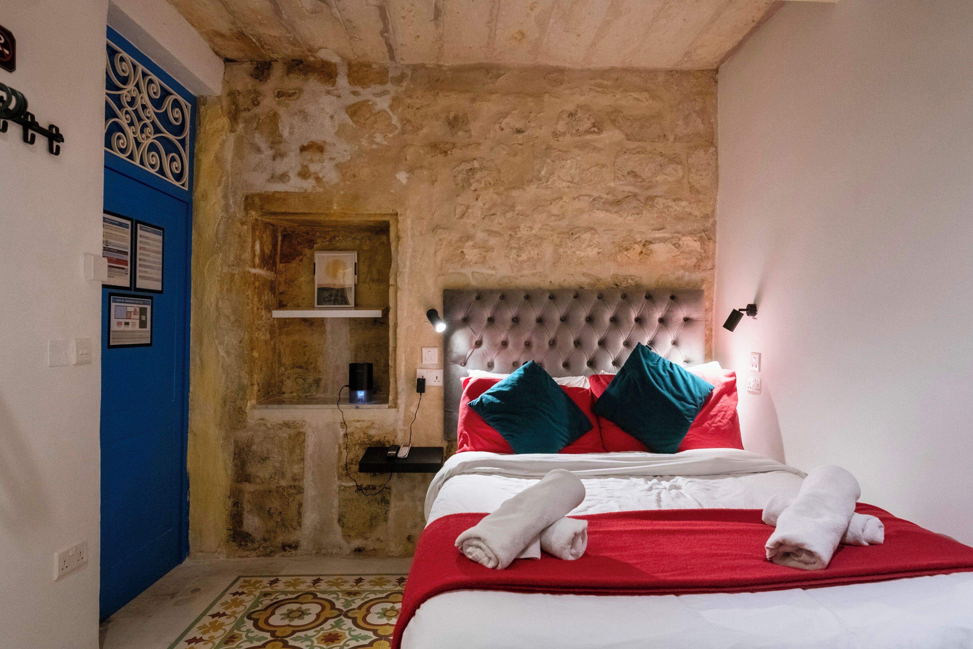 Un dormitorio con pared de piedra, una puerta azul, cama con ropa de cama roja y blanca, y cabecera con luces.