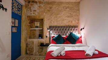 Un dormitorio con pared de piedra, una puerta azul, cama con ropa de cama roja y blanca, y cabecera con luces.