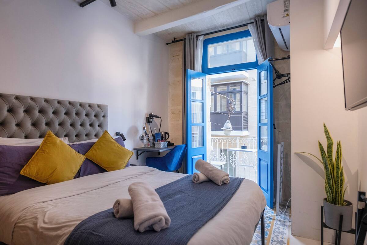 Un lit bien fait, doté d’une tête de lit capitonnée, une table de chevet avec une lampe, et une vue sur le balcon extérieur par une porte ouverte.