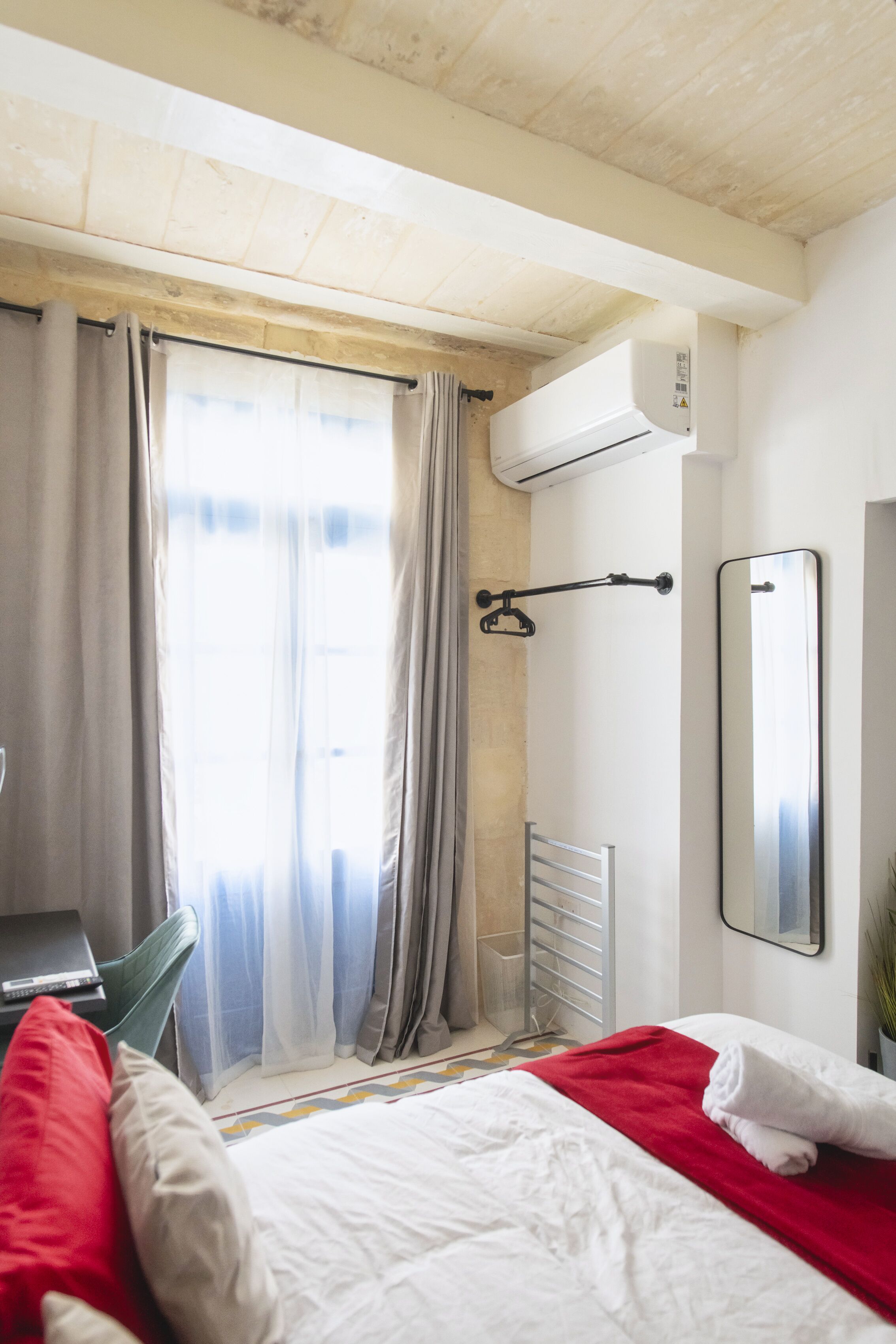 A hotel room with a bed, a desk, a chair, a radiator, and a window with curtains.