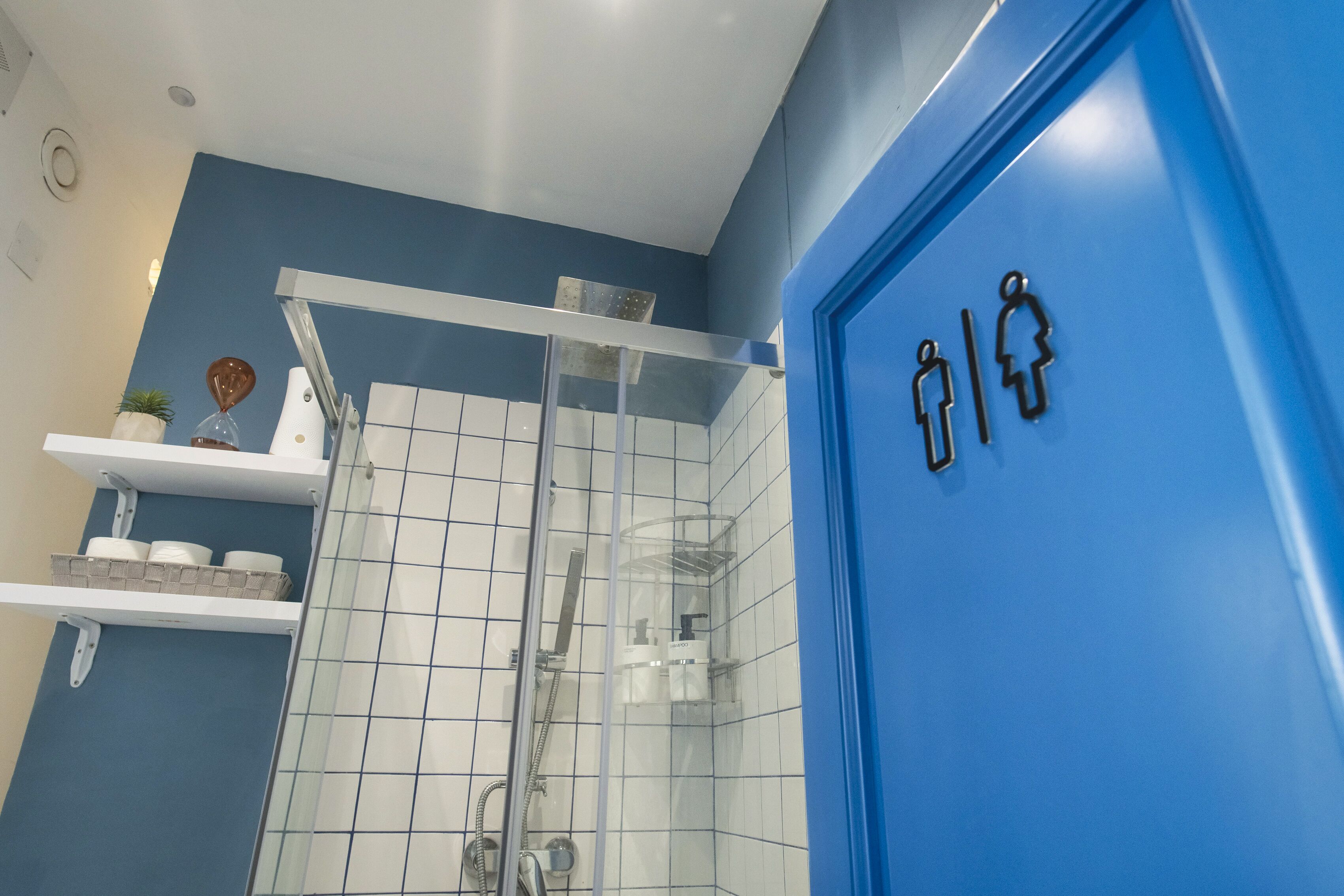 Ein Badezimmer mit blauer Wand, weißen Fliesen, einer Duschkabine aus Glas und einer blauen Tür mit Geschlechterzeichen.