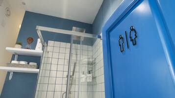 Ein Badezimmer mit blauer Wand, weißen Fliesen, einer Duschkabine aus Glas und einer blauen Tür mit Geschlechterzeichen.