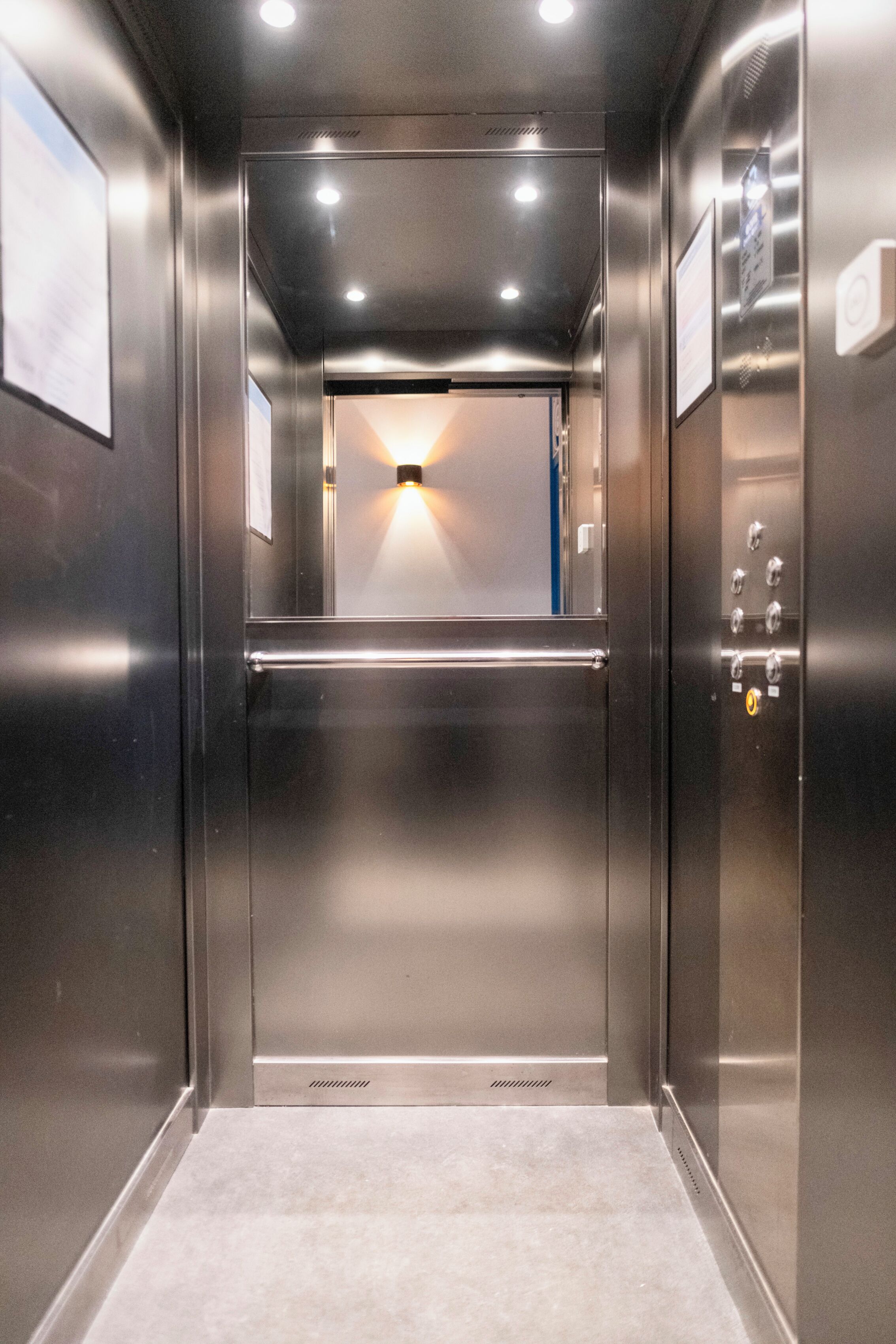 Interior de un ascensor con paredes metálicas, un pasamanos y una lámpara en la parte superior.