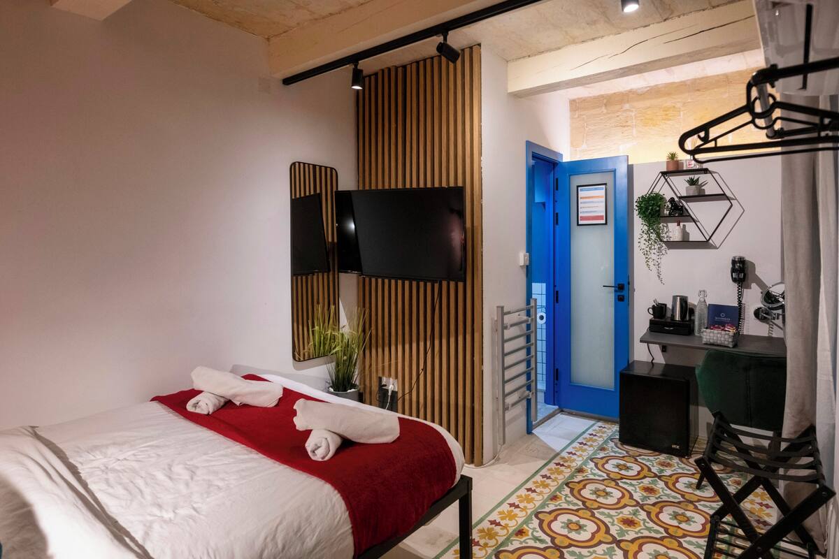 Une chambre d’hôtel moderne, équipée d’un lit, d’une télévision, d’un bureau avec une chaise et d’une salle de bain visible à travers une porte ouverte.