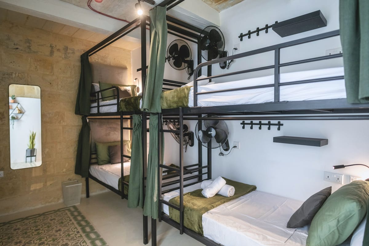 Une chambre avec un lit superposé, un rideau vert, une petite étagère et un ventilateur fixé au mur.