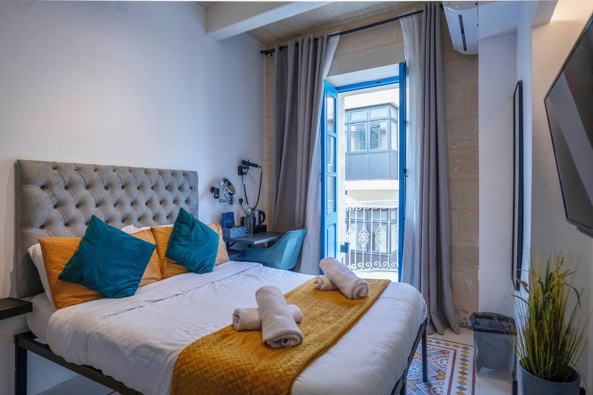 Un lit bien fait, doté d’une tête de lit capitonnée, d’un plaid jaune et de coussins bleus. On y trouve également un petit bureau avec une chaise et une plante en pot.