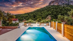 A rectangular swimming pool with lounge chairs and a wooden fence, surrounded by greenery and a mountainous backdrop. - Cozy apartment in Klek with swimming pool (Klek)