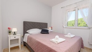 A bedroom with a bed, a nightstand, a lamp, a window with curtains, and a view of greenery outside. - Cozy apartment in Klek with swimming pool (Klek)