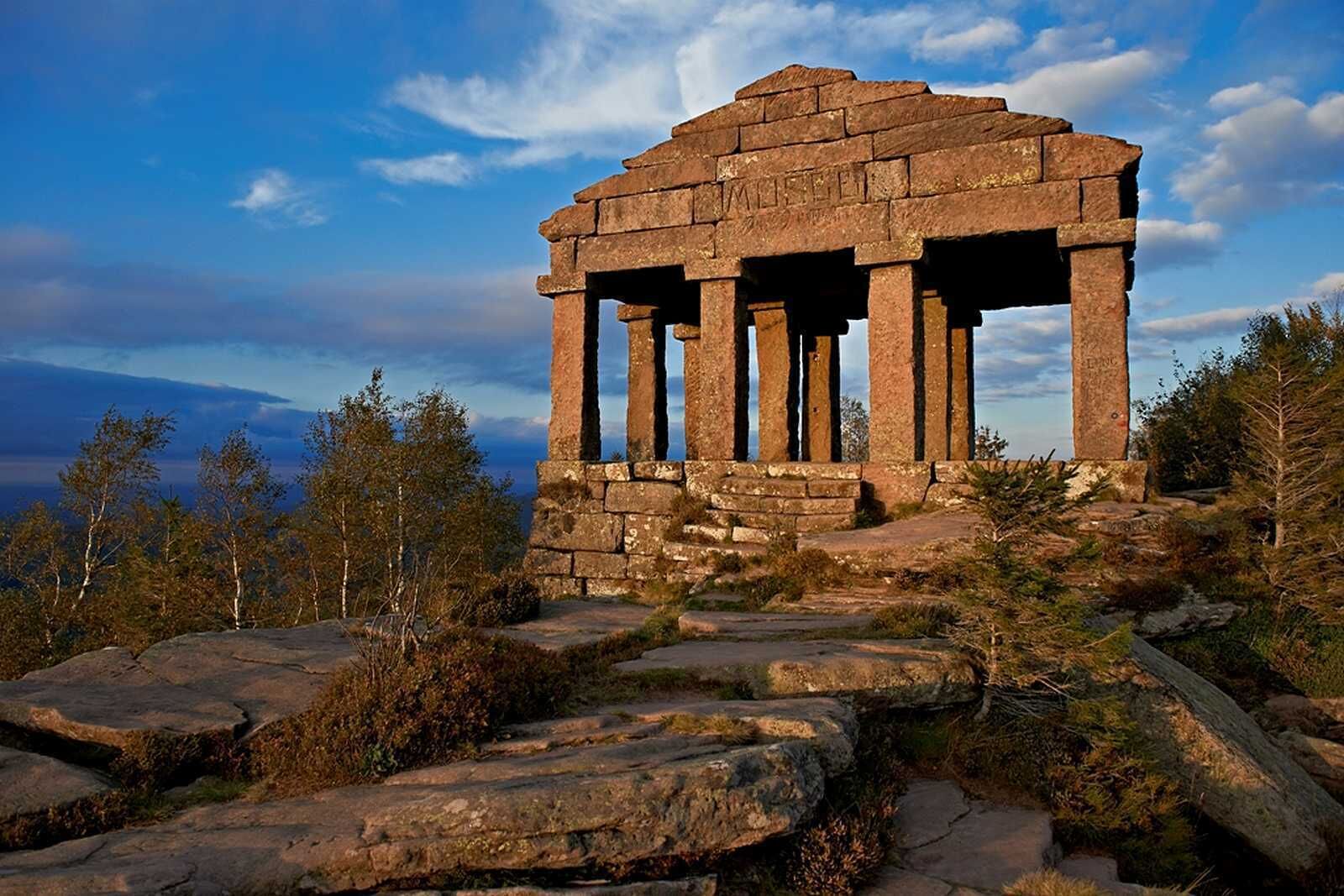 Una antigua estructura de piedra con columnas, rodeada de terreno rocoso y árboles.