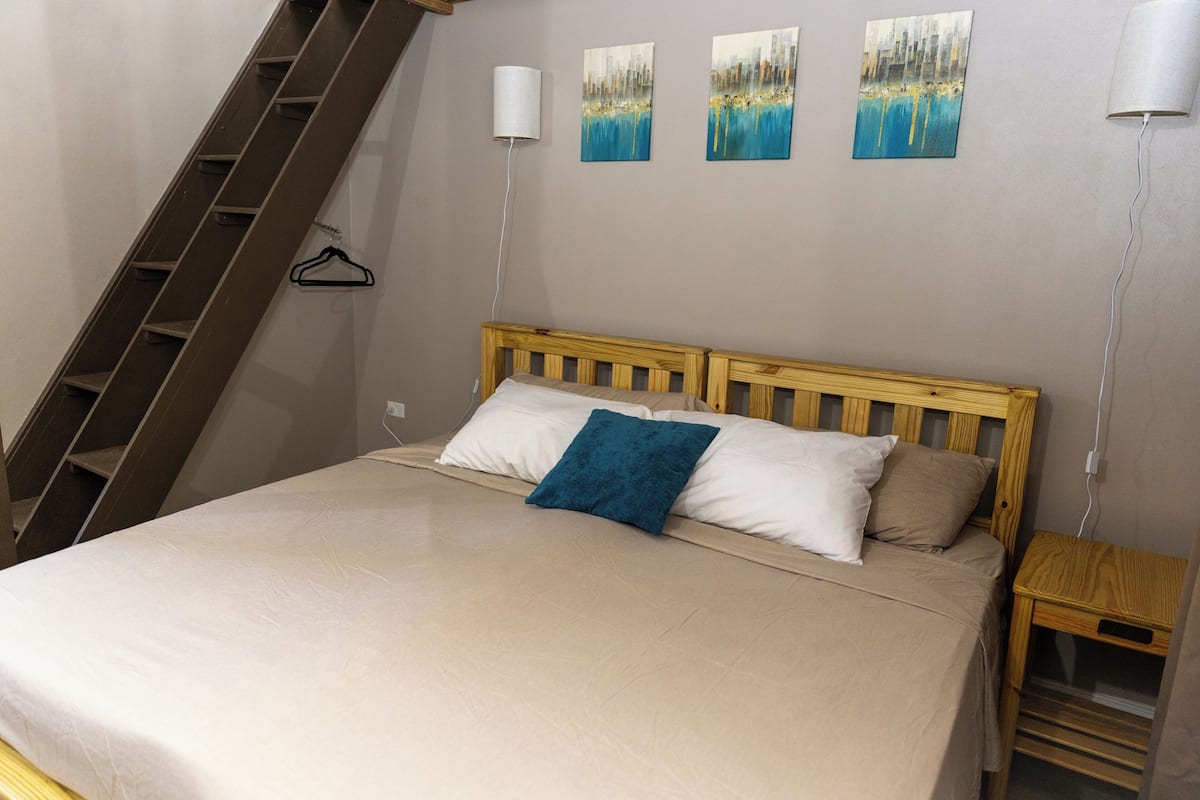A bedroom with a bed, a wooden headboard, a nightstand, a wall-mounted lamp, and a painting.