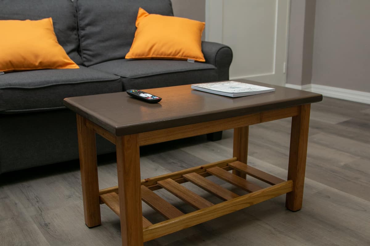 A wooden coffee table with a remote control and a magazine, placed in front of a grey sofa with orange cushions.