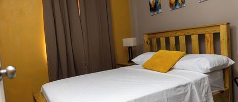 A bedroom with a wooden bed, white bedding, a yellow pillow, a nightstand with a lamp, and three paintings on the wall.