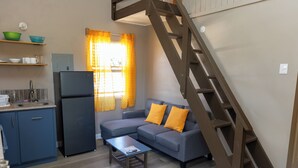 Un espace de vie compact comprenant un coin cuisine, un canapé avec des coussins orange, une table basse en bois et un escalier en bois.