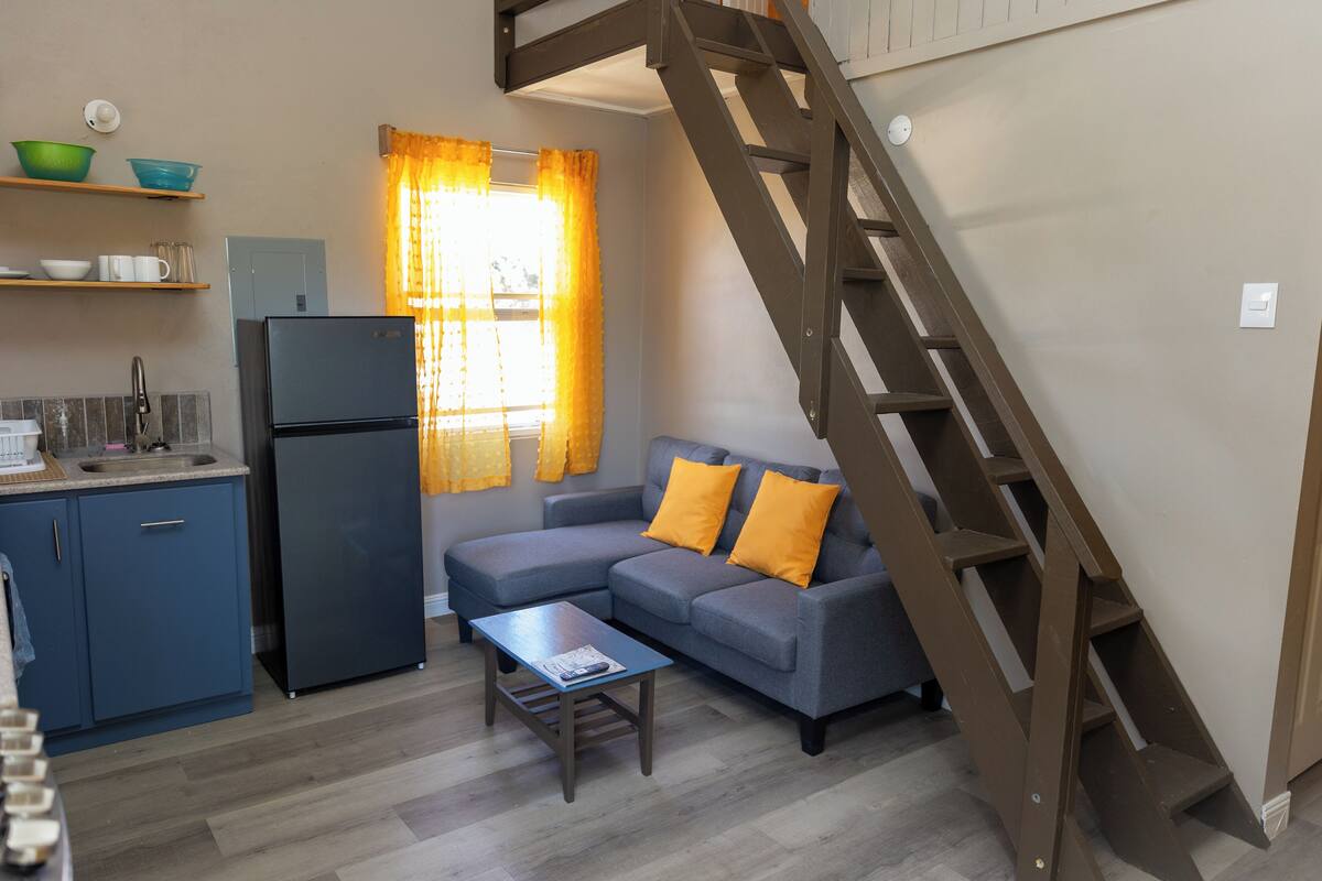 A compact living space with a kitchenette, a sofa with orange cushions, a wooden coffee table, and a wooden staircase.