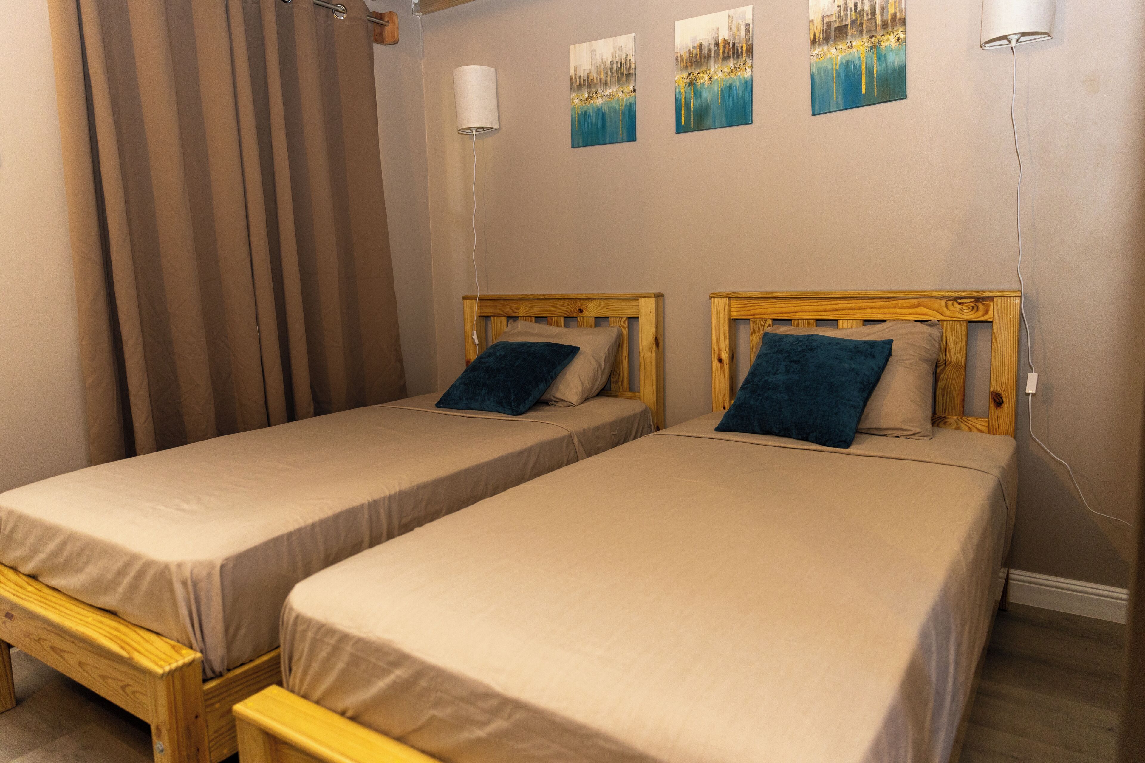 Dos camas individuales con cabeceros de madera y ropa de cama color beige, un banco de madera, una lámpara de pared y tres cuadros abstractos.