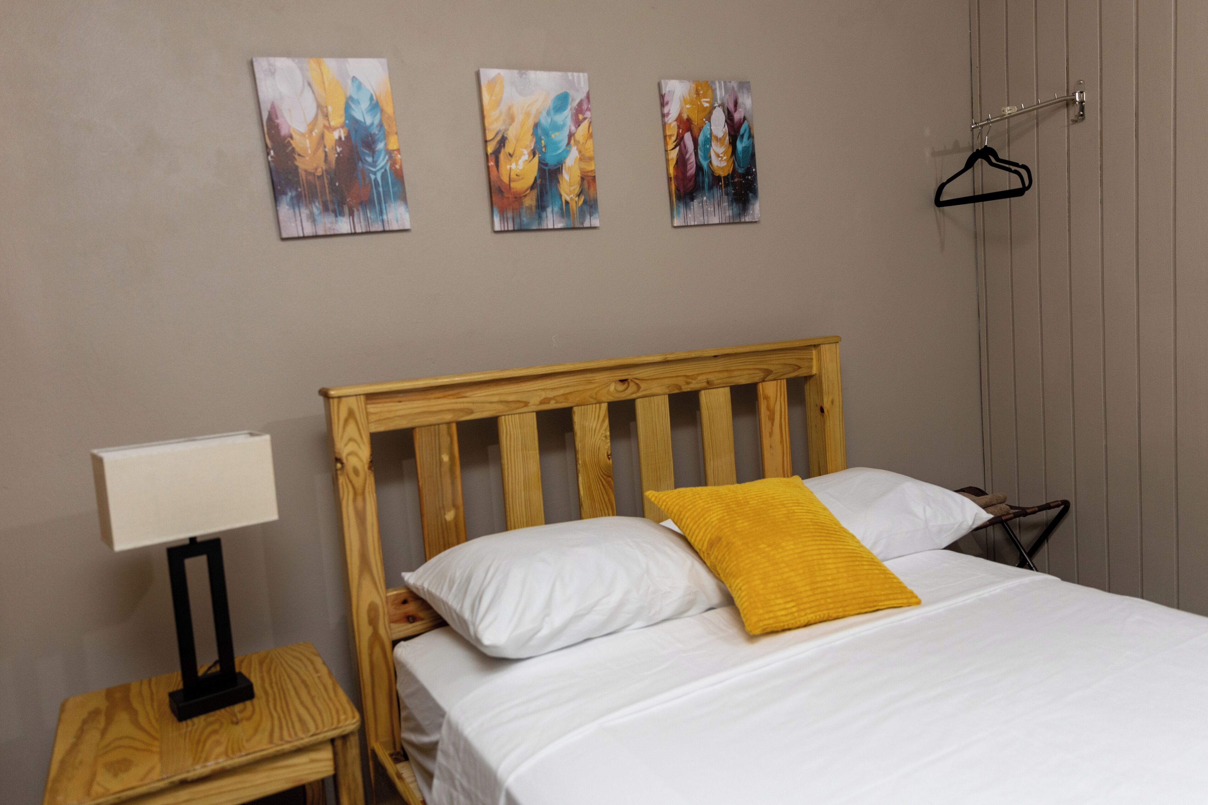 Un dormitorio con cabecera de madera, ropa de cama blanca, un cojín amarillo, una mesita de noche con lámpara y tres cuadros abstractos en la pared.