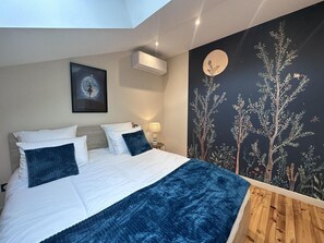Un dormitorio con una cama, una mesita de noche, aire acondicionado de pared, un cuadro enmarcado y un mural con árboles y una luna.