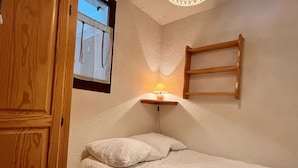 A small, neatly arranged bedroom with a single bed, a bedside table with a lamp, and a wooden shelf.