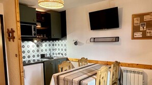 A compact kitchen with a small refrigerator, a sink, and a microwave. There is a dining table with chairs and a TV mounted on the wall.