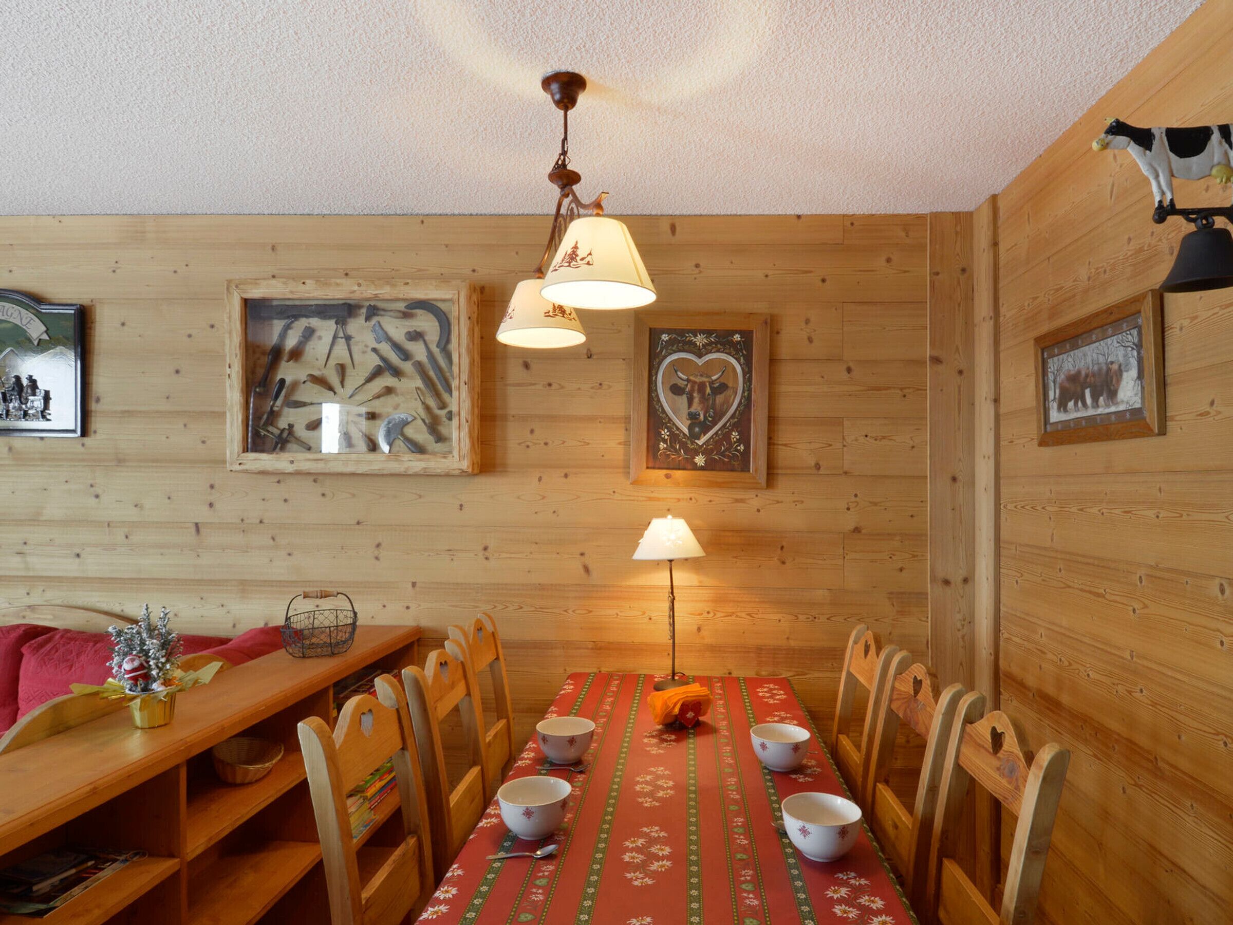A wooden dining room with a table set for four, two chairs on each side, a hanging lamp, and framed pictures on the walls.