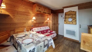 A cozy room with wooden walls, a dining table set with plates and glasses, a sofa with a patterned blanket, and a wall-mounted clock.
