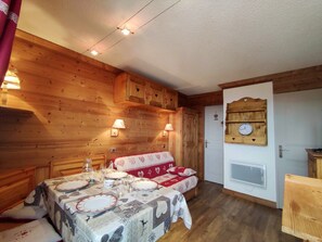Um quarto aconchegante com paredes de madeira, uma mesa de jantar posta com pratos e copos, um sofá com manta estampada e um relógio fixado na parede.