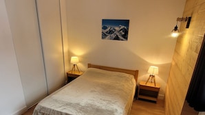 A single bed with a wooden frame and a grey bedspread in a room with wooden walls and floors. There are two bedside tables with lamps, and a framed picture of mountains on the wall. - Spacious apartment, mountain view - 7 guests (La Plagne)