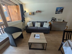 A cozy living room with a sofa, a coffee table, a chair, a bookshelf, and a kitchen area with a microwave. - Spacious apartment, mountain view - 7 guests (La Plagne)