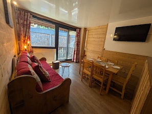 A cozy living room with a wooden sofa, a dining table with chairs, a flat-screen TV mounted on the wall, and large windows with curtains.