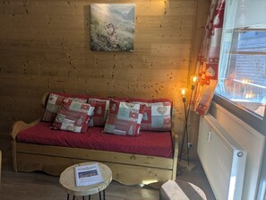 A cozy room with a wooden wall, a red sofa with patterned cushions, a small table with a magazine, and a window with curtains.