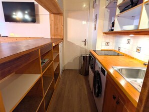 A compact kitchen with wooden shelves, a washing machine, a sink, and a television mounted on the wall.