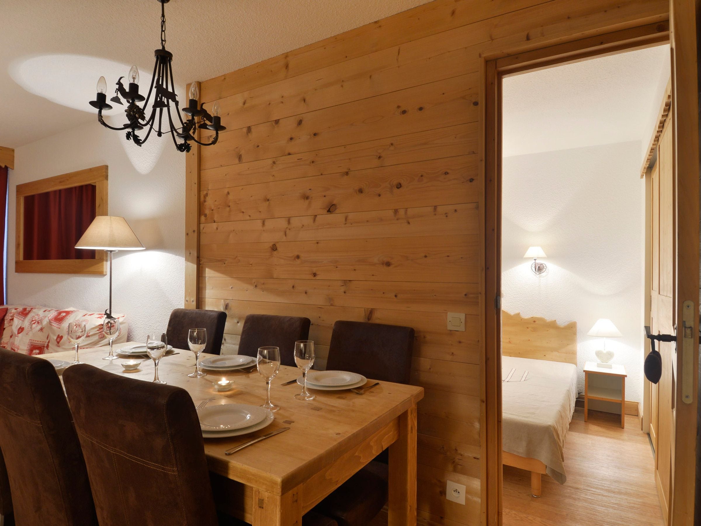 A wooden dining table with chairs, a chandelier, and a bed in the adjacent room.