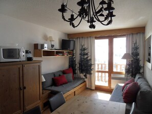 A cozy living room with a wooden table, a sofa, a television, and a ceiling light fixture. - Cozy ski-in/ski-out apartment - 6 guests (Belle Plagne)