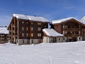 A multi-story wooden lodge with stone foundations, balconies, and a snow-covered roof, set against a clear sky.