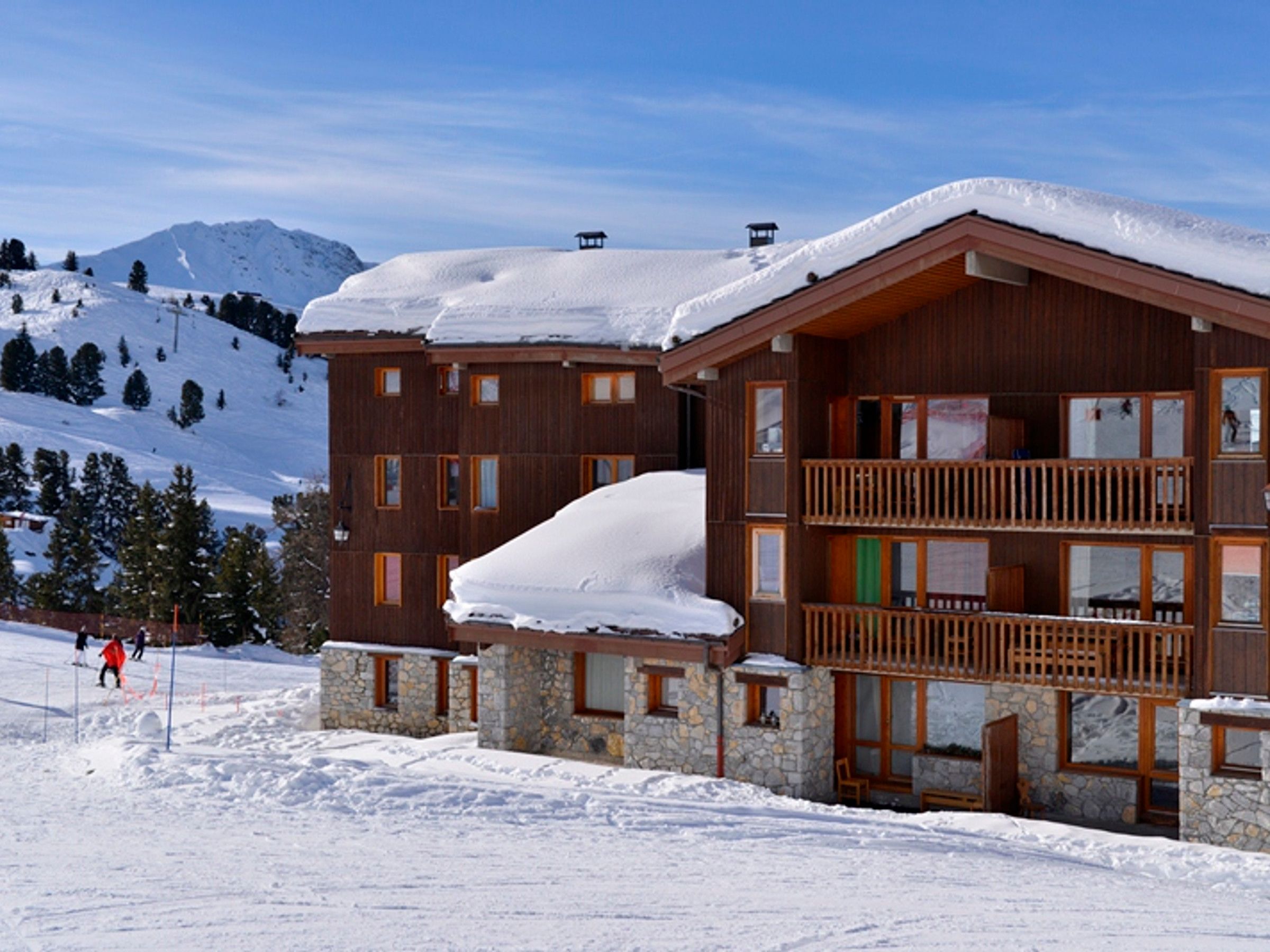 A multi-story wooden lodge with stone accents, a balcony, and a ski slope in the background.