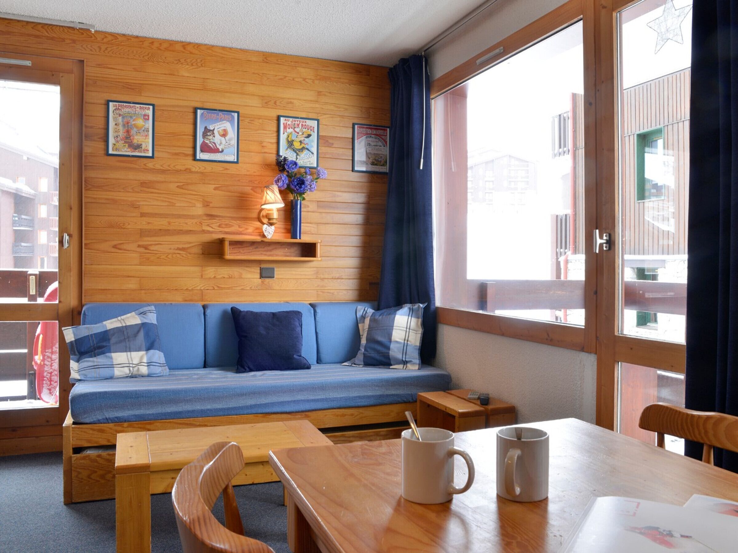 Ein gemütliches Zimmer mit einer Holzwand, einem blauen Sofa, einem Holztisch mit zwei Tassen und einem Fenster mit Vorhängen.