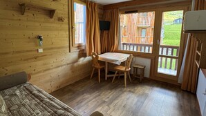A wooden cabin room with a bed, a table with chairs, a window with curtains, and a view of a wooden deck outside.