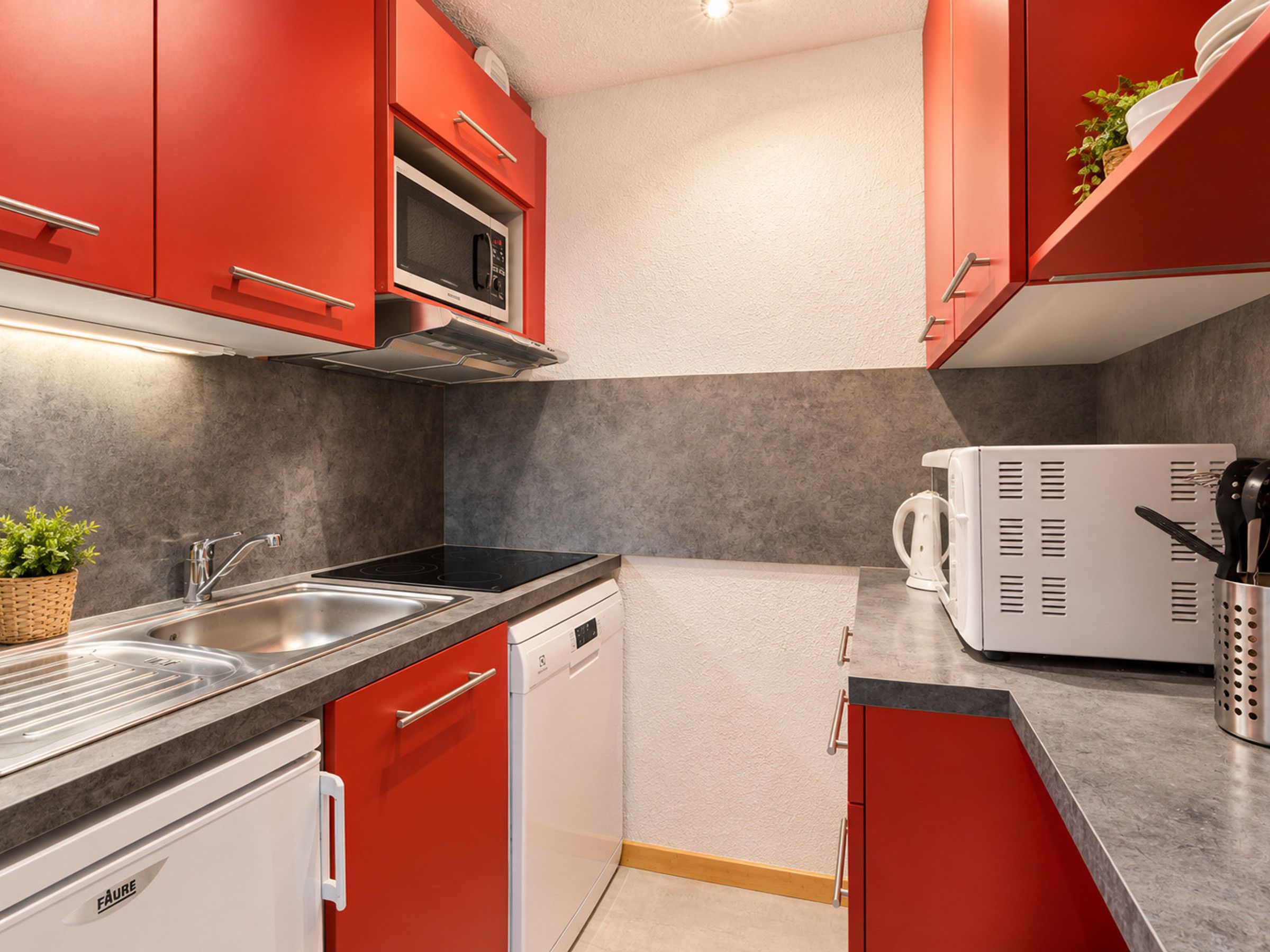 A compact kitchen with red cabinets, a built-in microwave, a coffee machine, and a dishwasher.