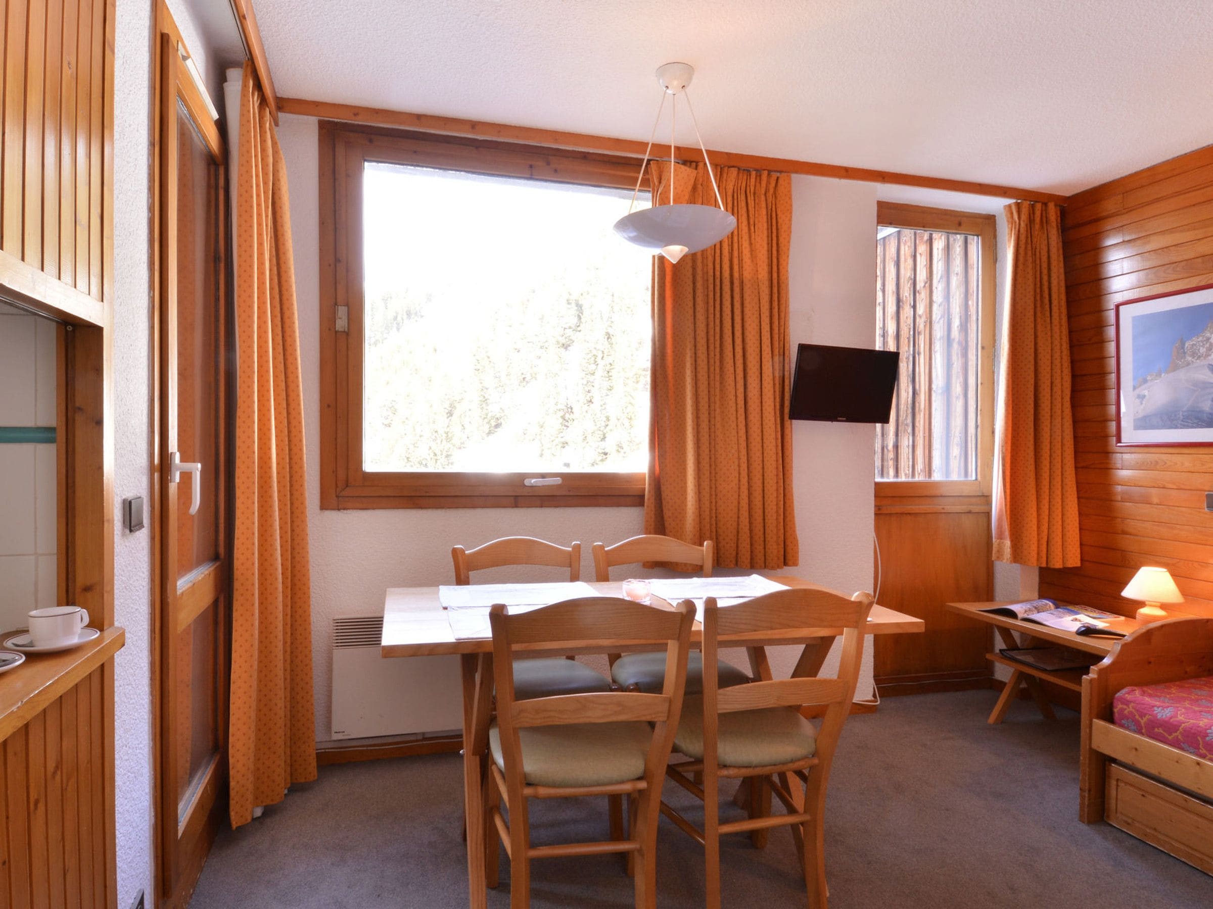 Une pièce de taille modeste, avec une table en bois, des chaises, un petit lit, un téléviseur et une fenêtre avec des rideaux.