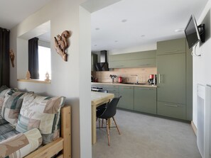 A compact living space with a sofa, a dining table with chairs, a kitchen area with green cabinets, and a mounted TV.