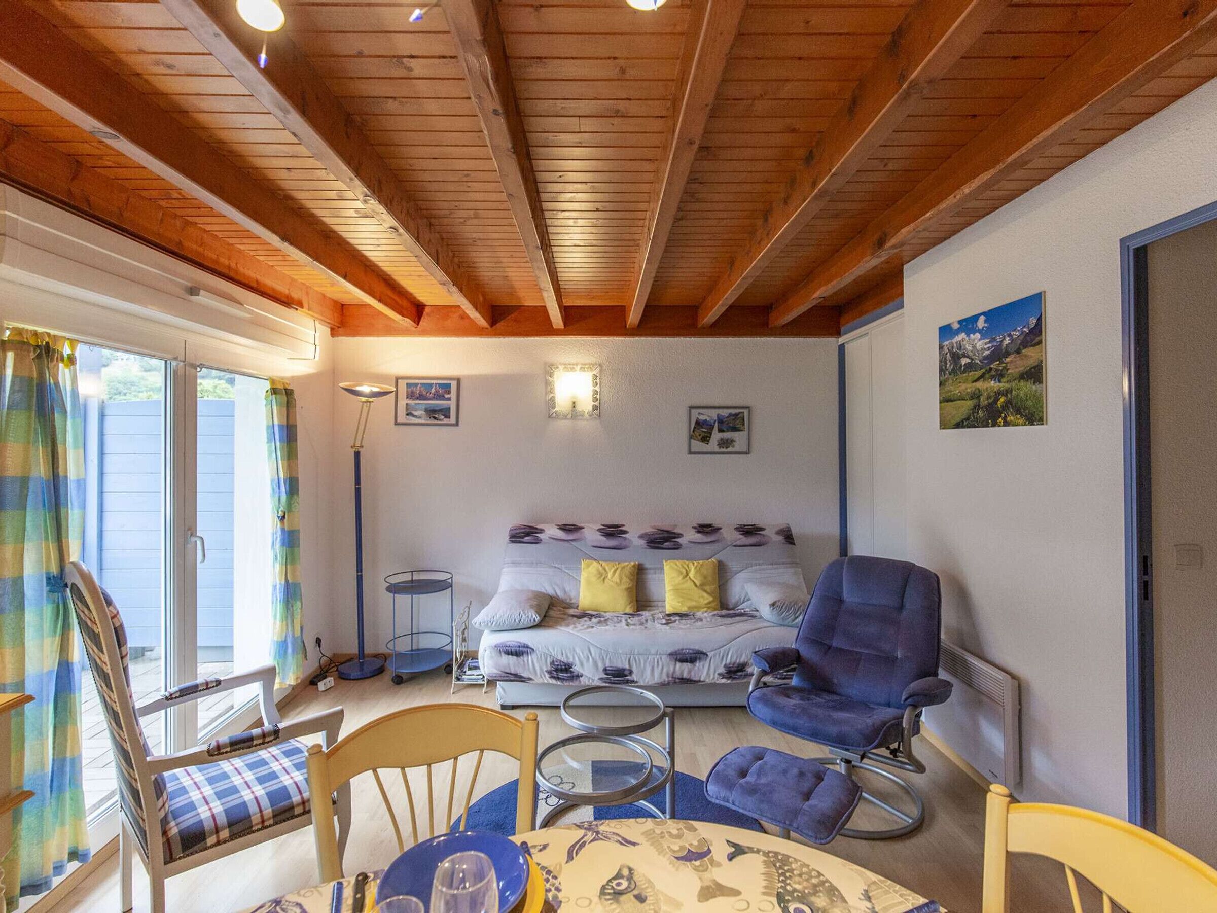 Una habitación acogedora con techo de madera, una cama con ropa de cama estampada, una silla azul, una mesita con camino de mesa y una lámpara de pie.