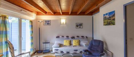 Una habitación acogedora con techo de madera, una cama con ropa de cama estampada, una silla azul, una mesita con camino de mesa y una lámpara de pie.