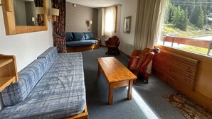 A cozy room with a sofa, a wooden table, and a chair. There is a window with a view of trees.
